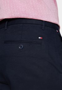 Pantaloni blu navy in tessuto intrecciato, con una tasca posteriore con chiusura a bottone e un piccolo logo rosso, bianco e blu.