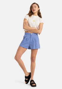 Un crop top blanc à côtes avec des bords festonnés et une broderie florale jaune, associé à un short plissé bleu et des sandales à plateforme noires.