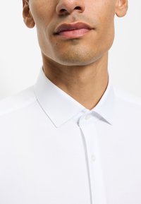 Homme portant une chemise blanche boutonnée au col, montrant le cou, la ligne de la mâchoire et les lèvres sur fond blanc.