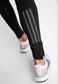 Leggings sportivi neri con tre strisce verticali grigie sul lato, dotati di caviglia con zip. Paio di scarpe da corsa grigie e blu visibili.