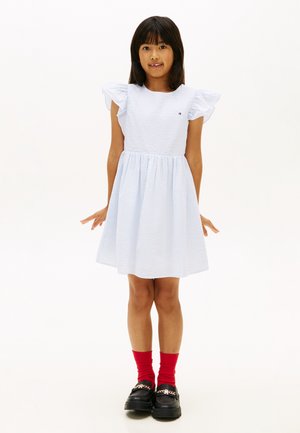 Młoda dziewczynka stojąca w białej teksturowanej sukience z falbanianymi rękawami, czerwonych skarpetkach i czarnych mokasynach z ozdobnym łańcuszkiem.