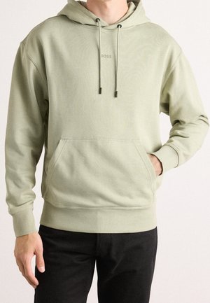 Homme portant un sweat à capuche vert clair avec poche avant et pantalon noir, main reposant dans la poche, fond neutre.