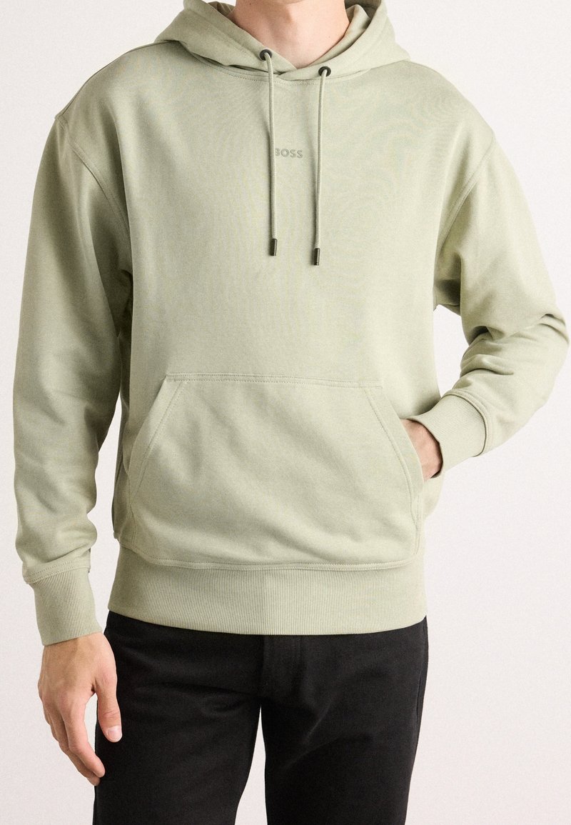 Homme portant un sweat à capuche vert clair avec poche avant et pantalon noir, main reposant dans la poche, fond neutre.