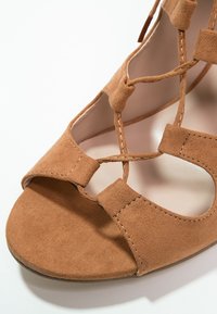 Sandale à talon en suède marron clair avec bout ouvert, brides croisées et design ajouré. Intérieur lisse avec détails de couture visibles.
