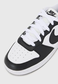 Nike sneakers med en overdel i sort læder og hvid syntetik, perforeret tå, hvide snørebånd og sort swoosh-logo på siden.