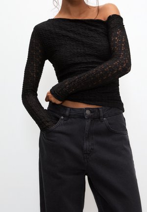 Femme portant un haut texturé noir à manches longues décolleté épaules et un jean noir taille haute, main rentrée dans la poche sur un fond uni.