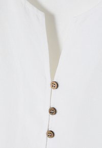 Tissu en lin blanc avec un décolleté en V et trois boutons en bois marron fixés verticalement au centre.