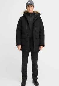 Crna parka s kapuljačom obloženom krznom, prednjim džepovima i zatvaranjem na patentni zatvarač, nosi se preko crnog topa i hlača, uparen s sivoj beanie.