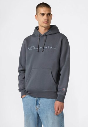 Grijze hoodie met een kangoeroezak, een trekkoord capuchon en geborduurd "Champion" logo. Voorzien van ribgebreide manchetten en zoom. Draagt samen met lichtblauwe jeans.