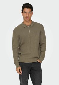 Õlivärvi ribakudumist pullover, millel on tõmblukk kaeluses, pikad varrukad ja nööbitud äär, koos tumedate teksadega.