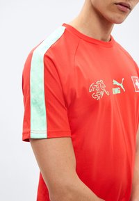 Άνδρας που φοράει κόκκινο αθλητικό πουκάμισο με κοντά μανίκια, με τιρκουάζ ρίγα στον ώμο και λευκά λογότυπα, συμπεριλαμβανομένου ενός λιονταριού και ενός εμβλήματος με σταυρό.