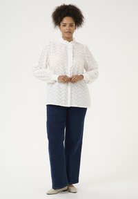 Camisa de botones blanca con patrón de zigzag y cuello con volantes, combinada con jeans de pierna recta azul oscuro y zapatos de lona color beige.