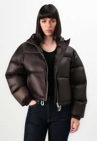 Giacca a vento marrone con finitura lucida, dotata di cappuccio, chiusura con zip e ampie sezioni trapuntate. Indossata sopra una maglietta nera e jeans blu.