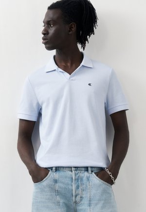 Uomo con dreadlocks che indossa una polo azzurro chiaro a maniche corte e jeans blu sbiaditi, in piedi con le mani in tasca, che guarda verso sinistra.