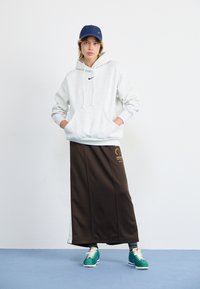 Grijze hoodie met zwart logo, bruine rok met gouden graphics, marineblauwe pet, groene sneakers met witte zolen, staand tegen een blauwe achtergrond.