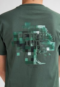 Zelené bavlněné tričko s grafickým designem stromu tvořeného pixelovanými zelenými čtverci, s výrazným potiskem "PETROL IND".