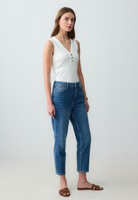 Top senza maniche bianco a coste con tre bottoni dorati sul collo, abbinato a jeans in denim blu a vita alta e sandali marroni con sabot.