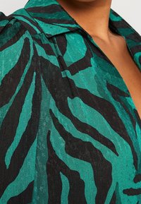 Gros plan d'une personne portant une chemise verte et noire à motifs avec un col et un tissu subtilement texturé. L'encolure est visible.