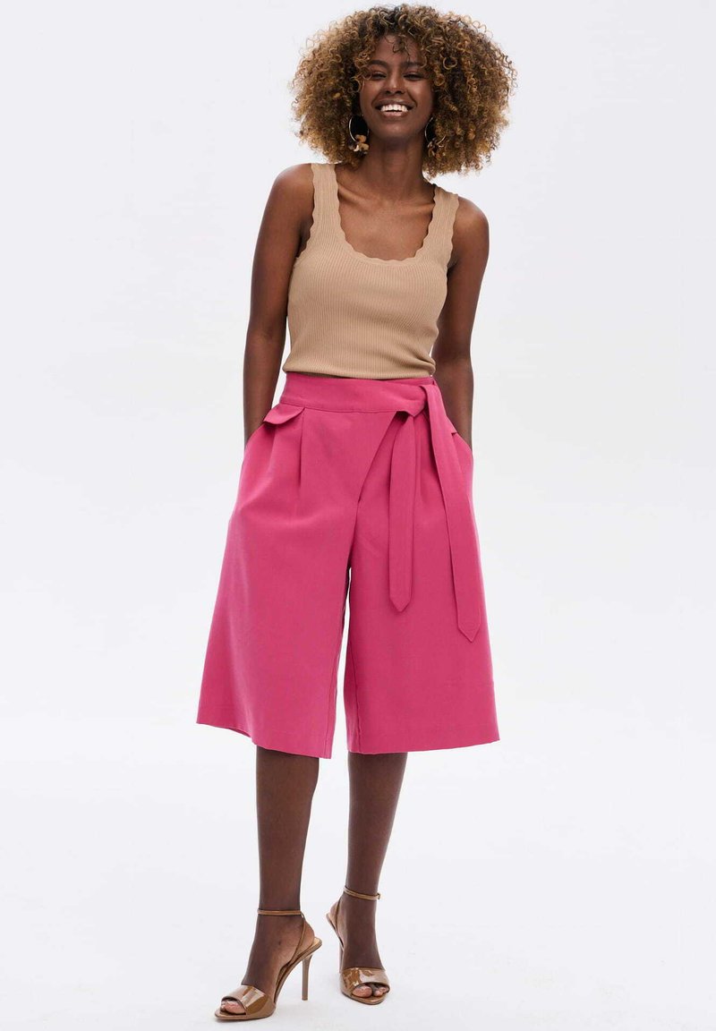 Roze wijde shorts met een knoopdetail in de taille, gecombineerd met een beige geribde tanktop; het model draagt bruine haksandalen.