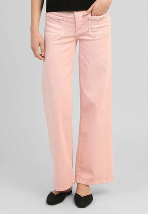Femme portant un pantalon large rose clair avec des poches plaquées à l'avant et des ballerines noires pointues, debout devant un fond gris.