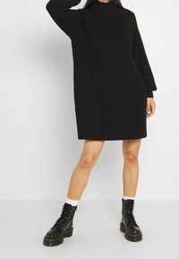 Robe en tricot noire avec un col haut, des manches longues et une coupe ample. Portée avec des bottes noires à lacets et des chaussettes blanches pour le contraste.
