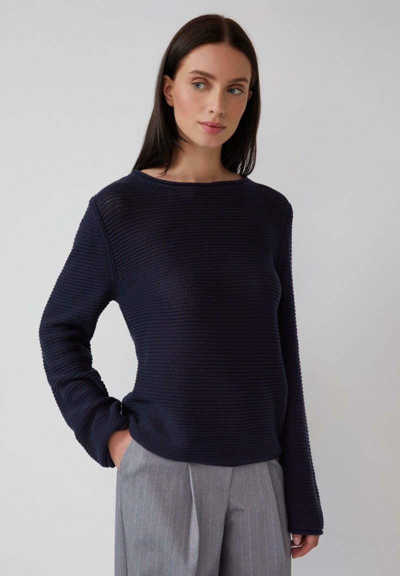 Frau mit dunklen Haaren, die einen strukturierten marineblauen Pullover und graue Nadelstreifenhosen trägt, steht mit einer Hand in der Tasche vor einem schlichten Hintergrund.