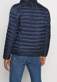 Marineblå pufferjakke med vandrette syninger, høj krave og sort logo-accent på skulderen, lavet af glat, skinnende materiale.