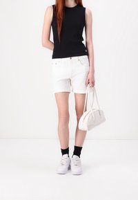 Svart ärmlös top, vita denimshorts, vita sneakers och en liten vit handväska. Svarta ankelsockor och en enkel silhuett.