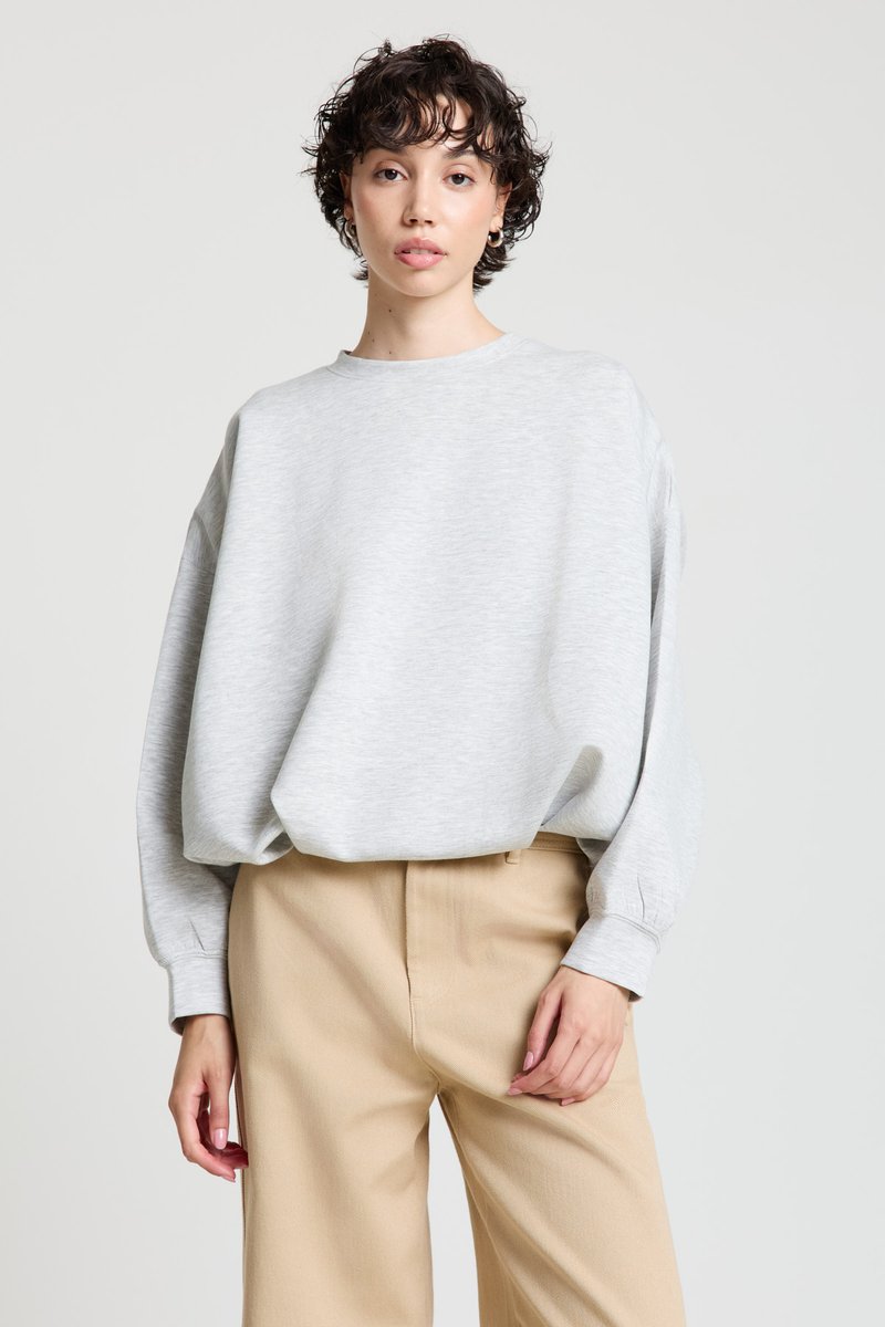 Felpa grigia chiara con scollo rotondo, spalle scese e maniche voluminose, abbinata a pantaloni beige con una vestibilità sartoriale.