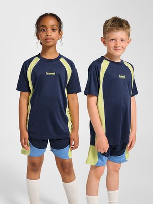 To børn står side om side iført marineblåt, lysegrønt og blåt sportstøj med hvide knæstrømper på en ensfarvet baggrund.