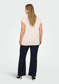 Haut beige clair à manches courtes avec une coupe décontractée, associé à un jean flare bleu foncé, porté avec des talons noirs, vu de dos.