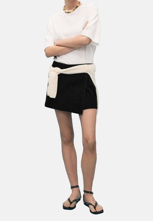 Persoon draagt een wit t-shirt, zwarte minirok met een crèmekleurige trui om de taille gebonden, zwarte gespte sandalen en een kralenketting.
