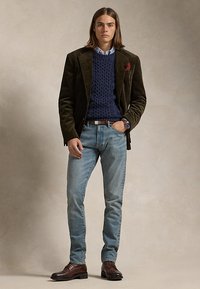 Bărbat tânăr purtând un sacou verde măsliniu închis, pulover albastru închis cu model torsadat, blugi albastru deschis, pantofi maro din piele și un ceas, stând în fața unui fundal neutru.