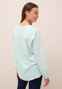 Mujer vistiendo una blusa de mangas largas a rayas verticales en azul claro y blanco, vista desde atrás, con pantalones oscuros sobre un fondo sencillo.