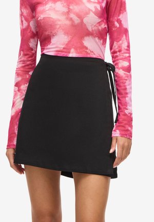 Žena nosící černou zavinovací sukni zavázanou na boku a růžovo-bílý dlouhý rukáv s batikovaným vzorem, stojící s rukama volně podél těla.