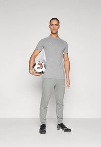 Tricou sportiv gri cu mâneci scurte și panou lateral alb. Pantaloni de trening gri. Pantofi sport negri. Ținând o minge de fotbal alb-negru.