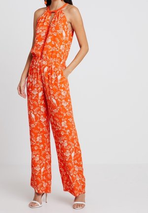 Vrouw draagt een mouwloze oranje jumpsuit met witte bloem- en vogelpatronen, gecombineerd met witte sandalen met hak.