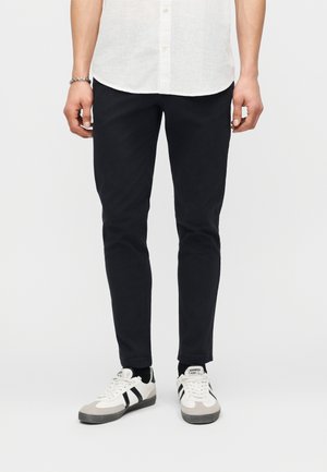 Férfi vékony fekete nadrágban, fehér ingben, fekete zokniban és fekete csíkos fehér tornacipőben, világos háttéren állva.