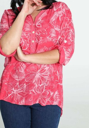 Femme portant un chemisier à boutons à fleurs rose vif avec les manches retroussées et un jean bleu foncé, les bras croisés et la main près du visage.