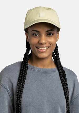 Joven mujer con largas trenzas negras que lleva una gorra beige y una sudadera gris, sonriendo contra un fondo claro y liso.