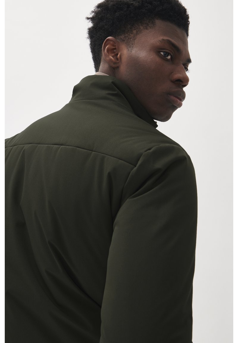 Veste vert foncé avec un col haut, texture lisse et design rembourré. Présente des coutures dans le dos et au niveau des épaules.
