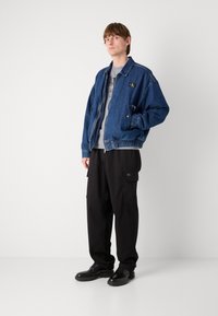 Veste en denim bleu foncé, avec une fermeture éclair et un ourlet élastiqué, associée à un pantalon cargo noir et des chaussures à lacets noires.