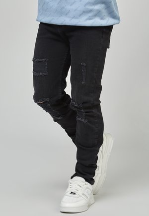 Zwarte distressed slim-fit jeans met rafelige patches en gescheurde details, gecombineerd met witte sneakers met een textuur.