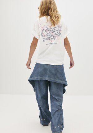 Personne avec des cheveux blonds s'éloignant, portant une chemise blanche à manches retroussées avec le texte "Créer de la magie" et un pantalon large à rayures bleues.