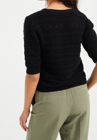 Maglione nero in tessuto strutturato con un motivo geometrico e sezioni a maglia aperta. Polsini e orlo a coste, abbinato a pantaloni verde chiaro.