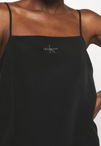 Schwarzes ärmelloses Oberteil aus glattem Stoff, mit dünnen Trägern und einem kleinen, reflektierenden Calvin Klein Jeans-Logo auf der Vorderseite.