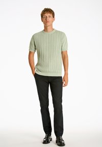 Lyhythihainen, vaaleanvihreä neulepusero, jossa on palmikkokuvio, yhdistettynä mustiin housuihin ja kiiltäviin mustiin kenkiin. Yksinkertainen, vartalonmyötäinen malli.