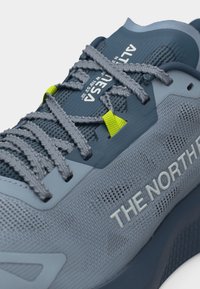 Κοντινό πλάνο αθλητικού παπουτσιού The North Face σε μπλε-γκρι χρώμα, με υφασμάτινη υφή, γκρίζα κορδόνια με κίτρινους μηχανισμούς στερέωσης και λεπτομέρειες στη γλώσσα με το λογότυπο της εταιρείας.