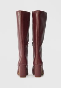 Μπουνέλες από burgundy δέρμα μέχρι το γόνατο με λεία υφή, τετράγωνα τακούνια και πίσω φερμουάρ, προσαρμοσμένες με κατακόρυφες ραφές για δομή.