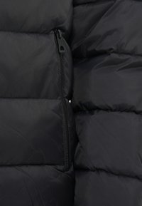 Μαύρος πούφος μπουφάν από ύφασμα με ραμμένες περιοχές, που διαθέτει τσέπη με φερμουάρ στην αριστερή πλευρά. Λείο, ελαφρύ υλικό με ματ φινίρισμα.